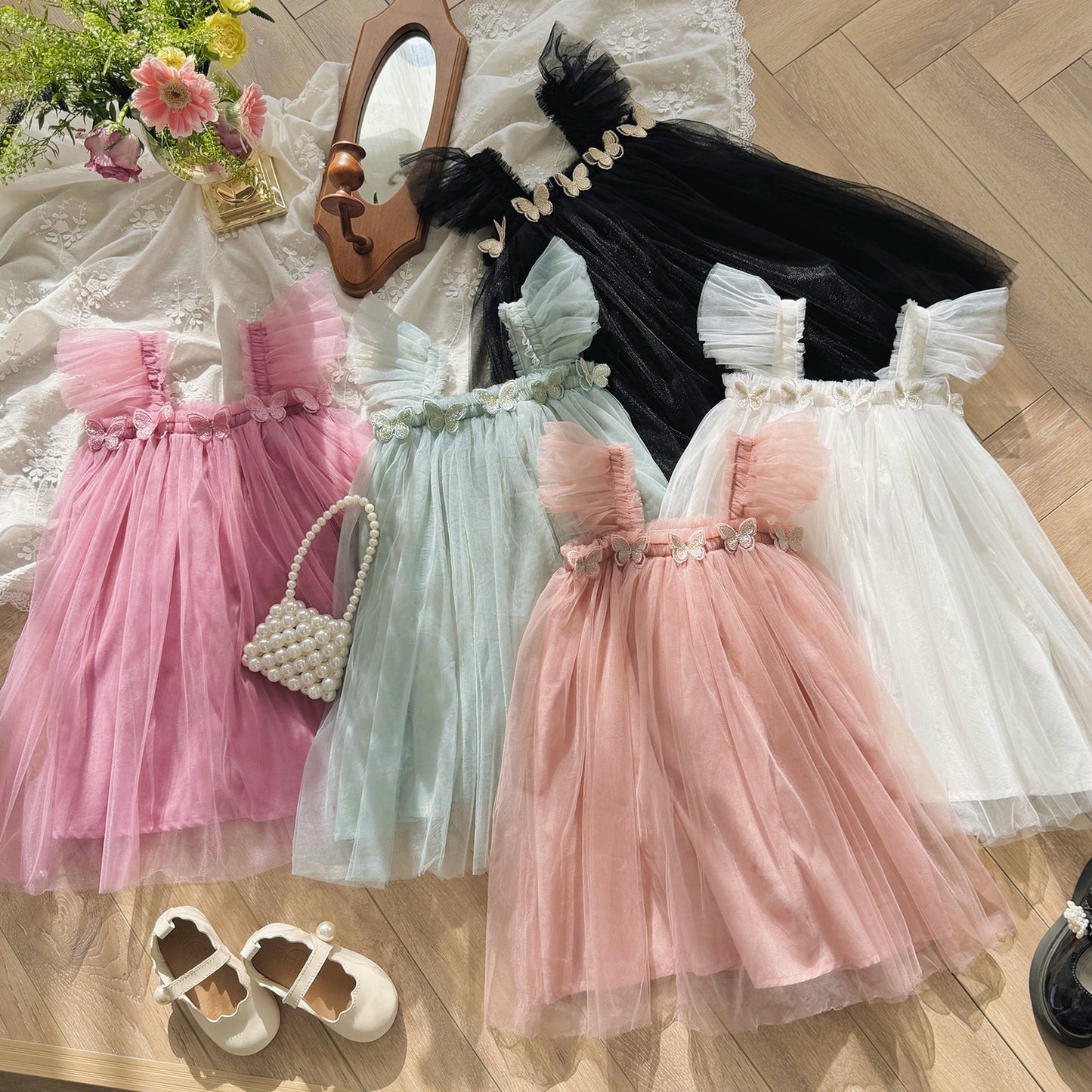 Four children's dresses in pink, green, and white on a wooden floor with a mirror and flowers.